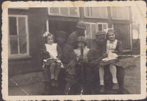 Foto van de kinderen van de familie Van de Vossenberg met Afro-Amerikaanse soldaten, gemaakt voor hun oude kippenschuur tijdens de Tweede Wereldoorlog (Foto: Privécollectie Wim van de Vossenberg, oktober 1944)